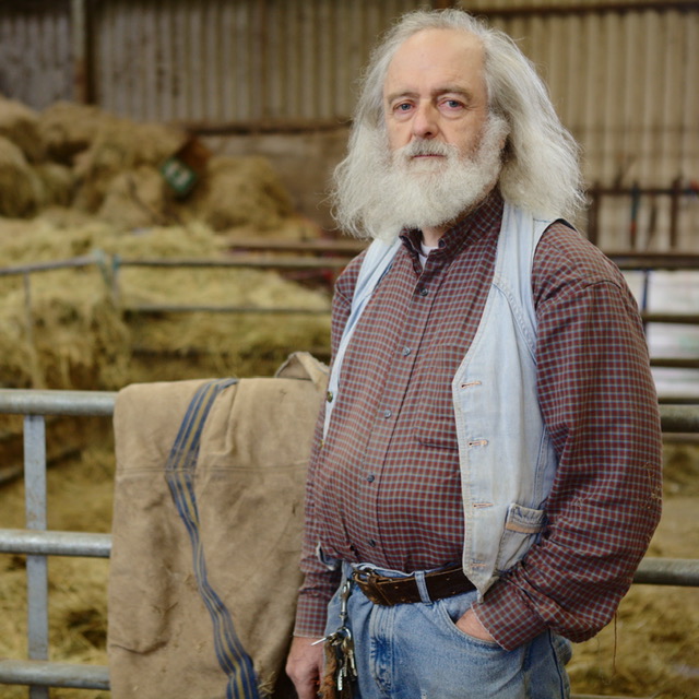 Charles Bishop, a man with long grey hair and full white beard, looking into the camera. the background, a barn with hay, is out of focus.
