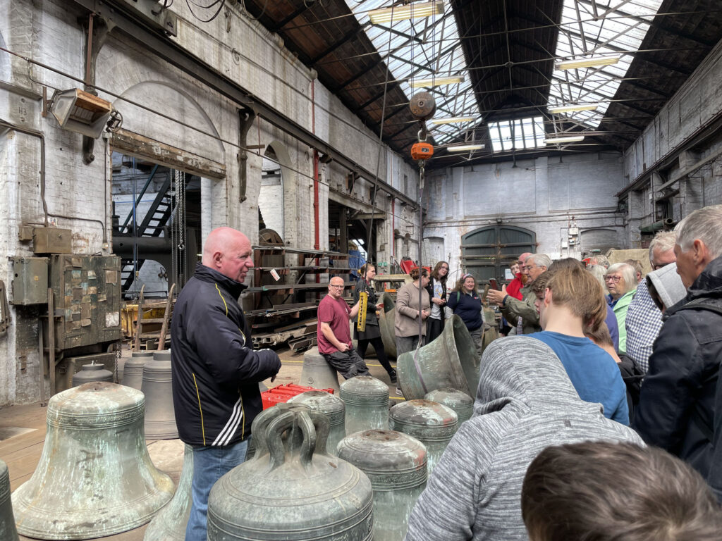 Internal view of large industrial building. Old and new bells sitting on the floor. People listening to a talk.